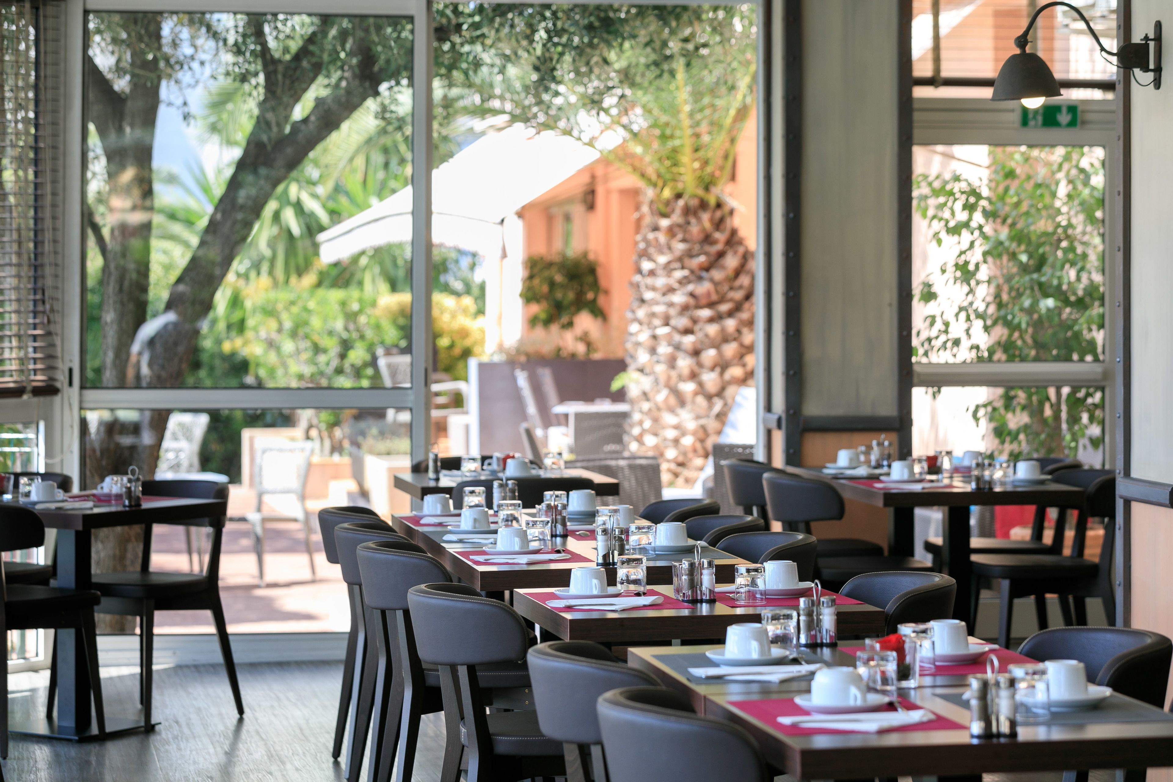 A restaurant with tables set for dining, large windows with a view of palm trees and a building, and a modern interior design.