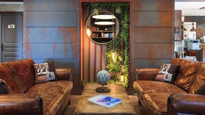 Una habitación con dos sofás de cuero, una mesa de centro de madera con un globo terráqueo y una revista, una estantería con luces colgantes y una pared verde con plantas.