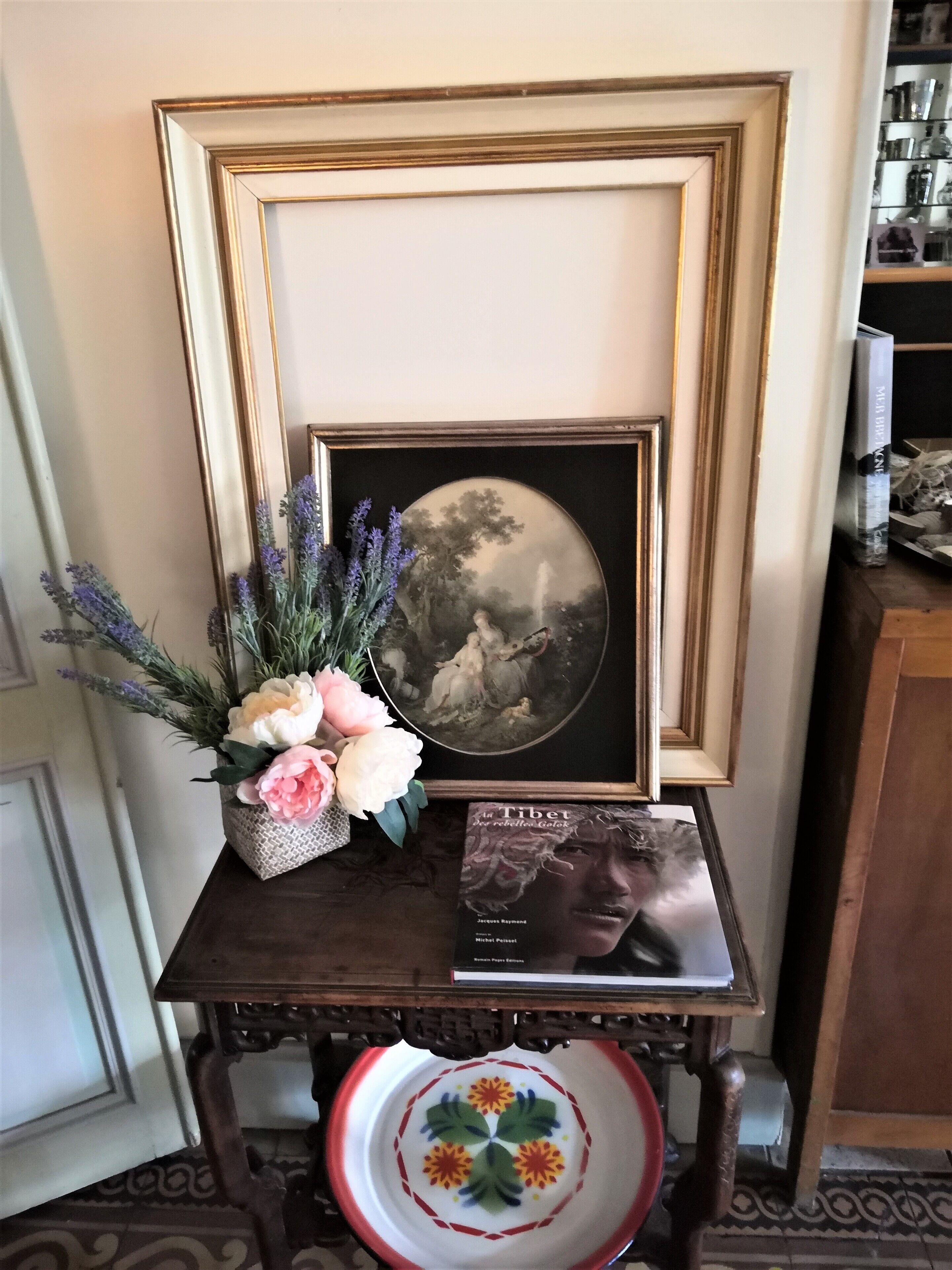 A framed painting on a dark wooden table, a vase of flowers, and a book titled "La Tibet".
