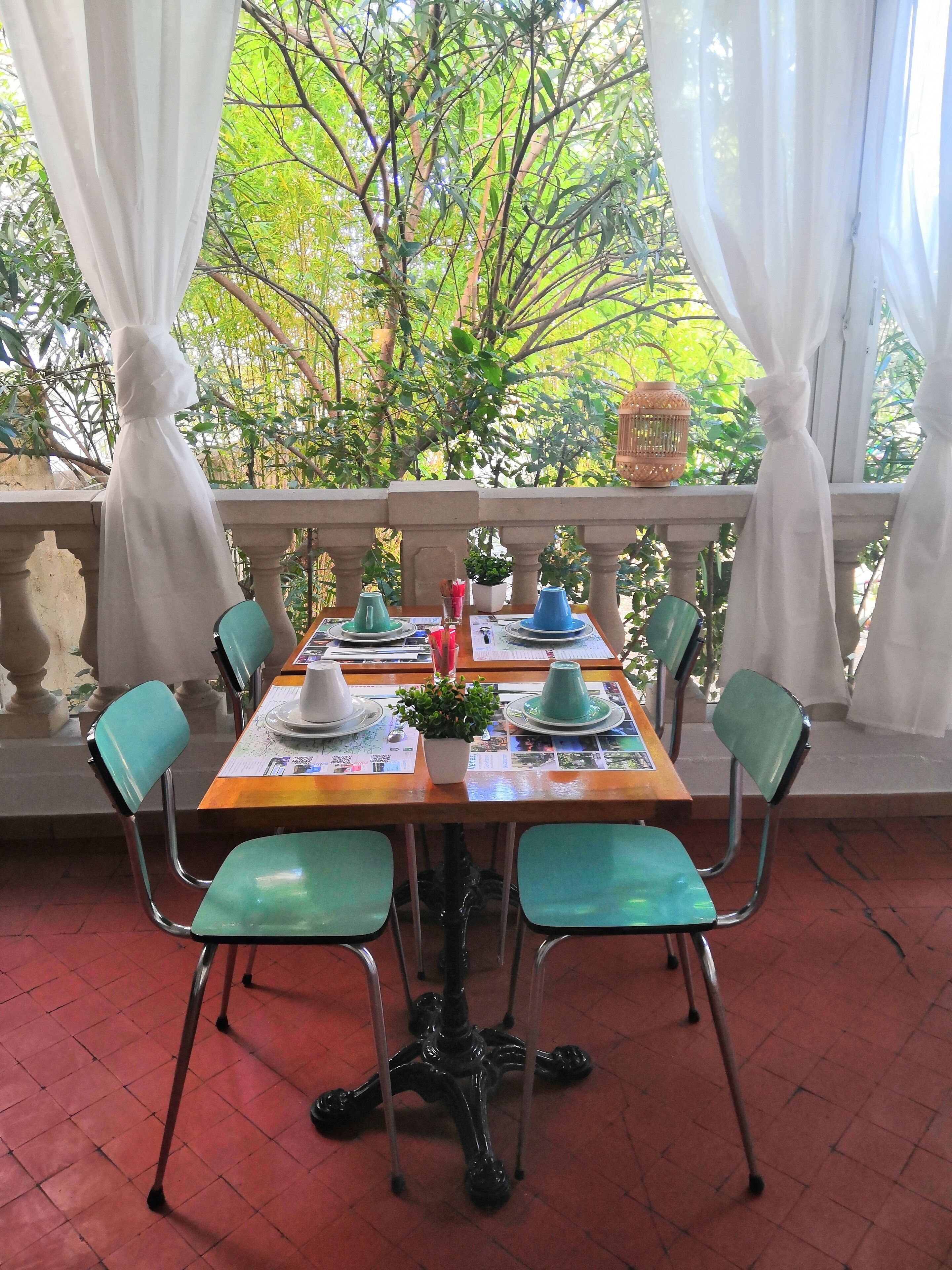Mesa de comedor dispuesta con platos, tazas y una planta en maceta, rodeada de sillas con estructura metálica y cojines verdes, en un balcón con vistas a árboles y una barandilla.