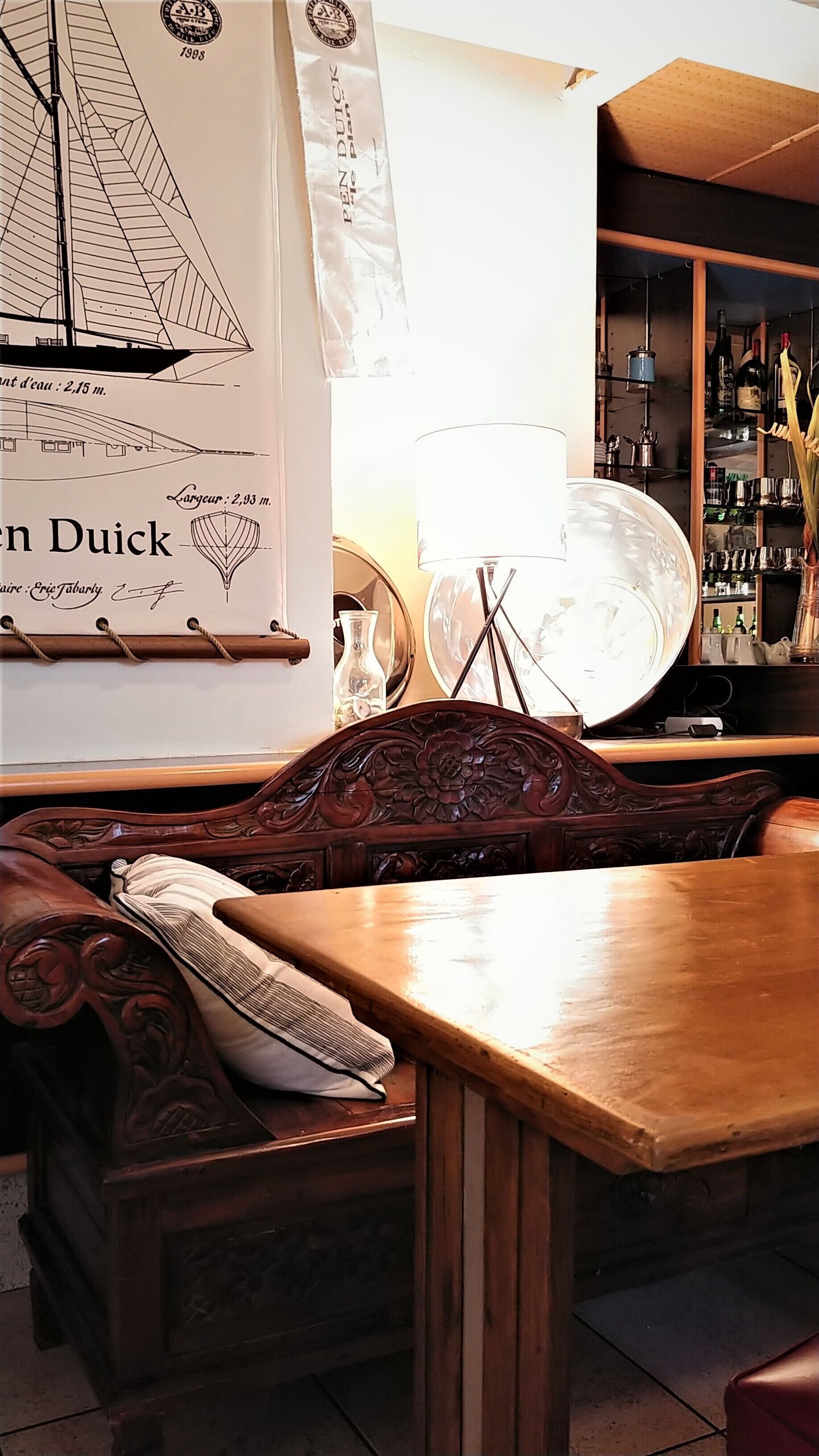 Une table et un banc en bois se trouvent dans une pièce où l’on aperçoit une grande affiche représentant un voilier, une lampe et une étagère remplie de bouteilles.