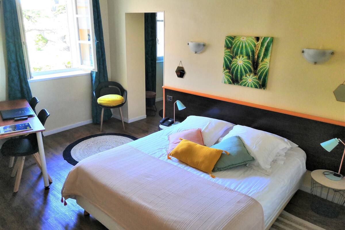 Ein Hotelzimmer mit Bett, Schreibtisch, Stuhl, Lampe und einer Wandmalerei von einem Kaktus.