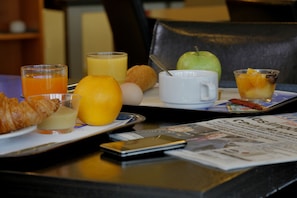 A breakfast tray with croissant, orange juice, a glass of orange juice, a green apple, a muffin, and a cup of coffee on a table with a newspaper and a smartphone.