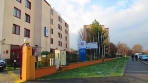 A hotel with a modern facade, a fenced parking area, and a clear sky. - Hotel Gallieni (Gennevilliers)