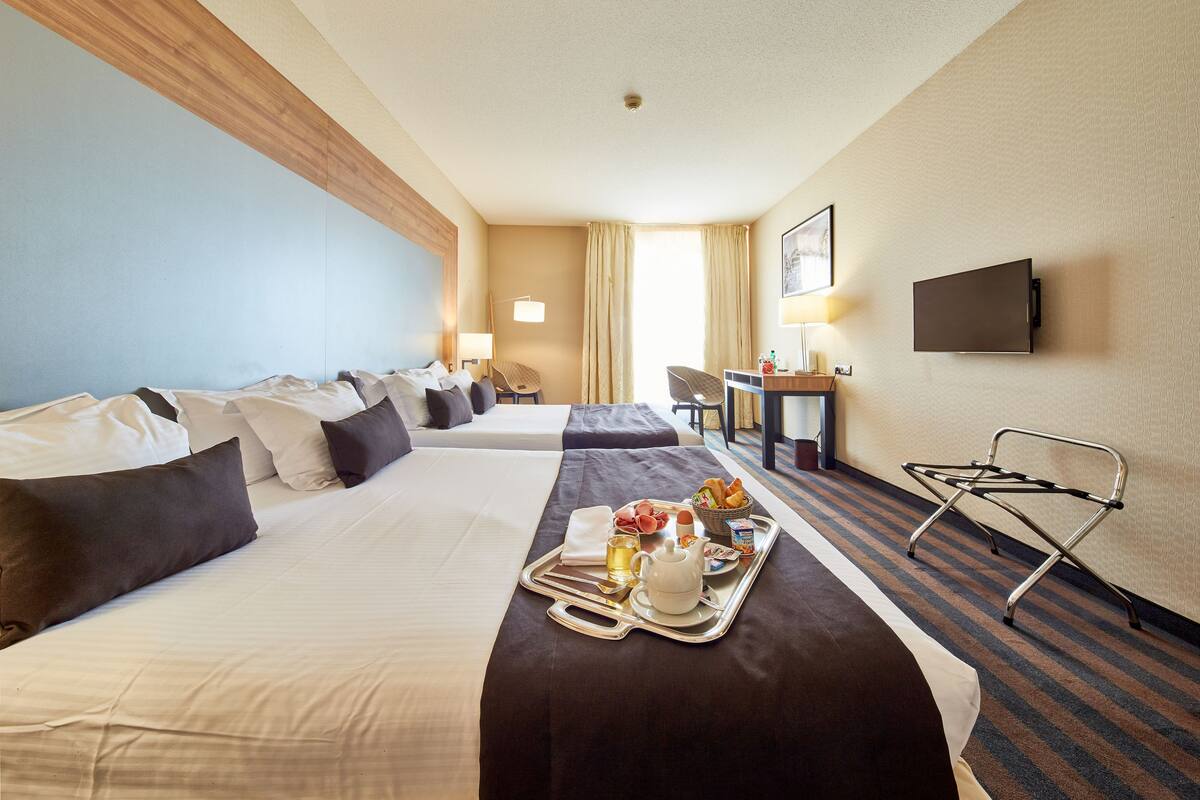 A hotel room with two beds, a desk, a chair, a TV, and a tray with breakfast items.