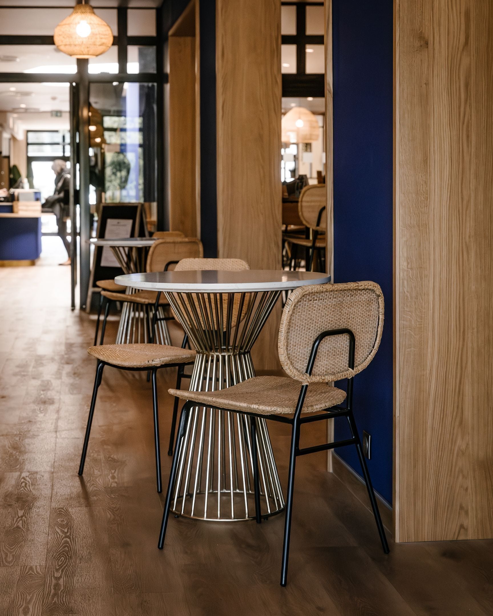 Un espace repas moderne avec des tables rondes et des chaises en osier, un plancher en bois et un mur d’accent bleu.