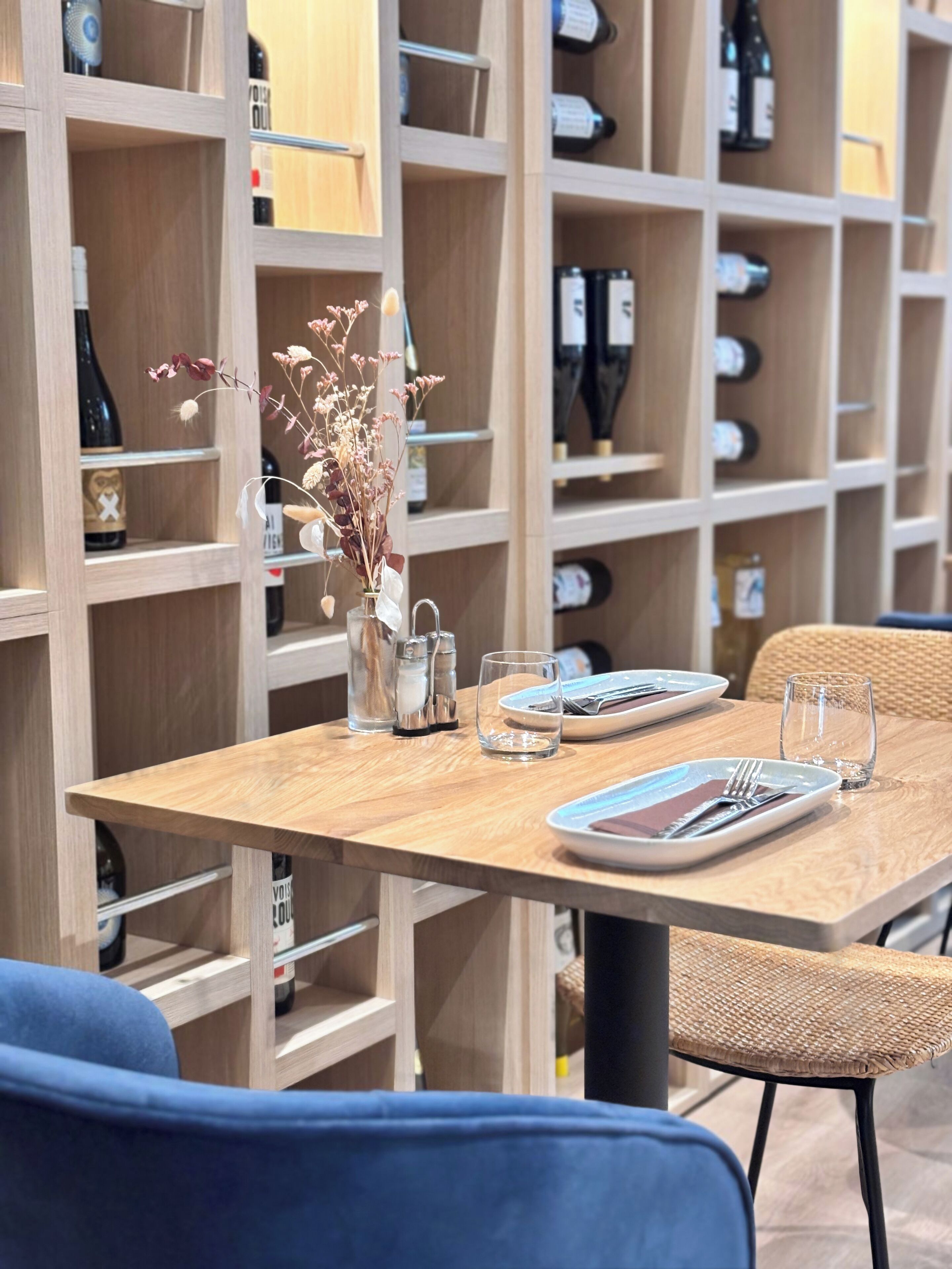 Un comedor con una mesa de madera dispuesta para dos personas, un florero con flores secas, saleros y pimenteros, y botellas de vino en estantes al fondo.