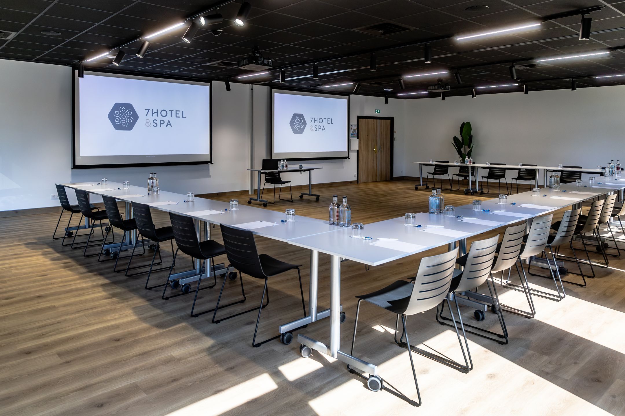 A conference room with a long table, chairs, a projector screen displaying '7HOTEL & SPA', and a wooden floor.