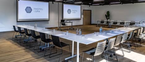 Un salón de conferencias con una larga mesa, sillas, una pantalla de proyector que muestra '7HOTEL & SPA' y piso de madera.