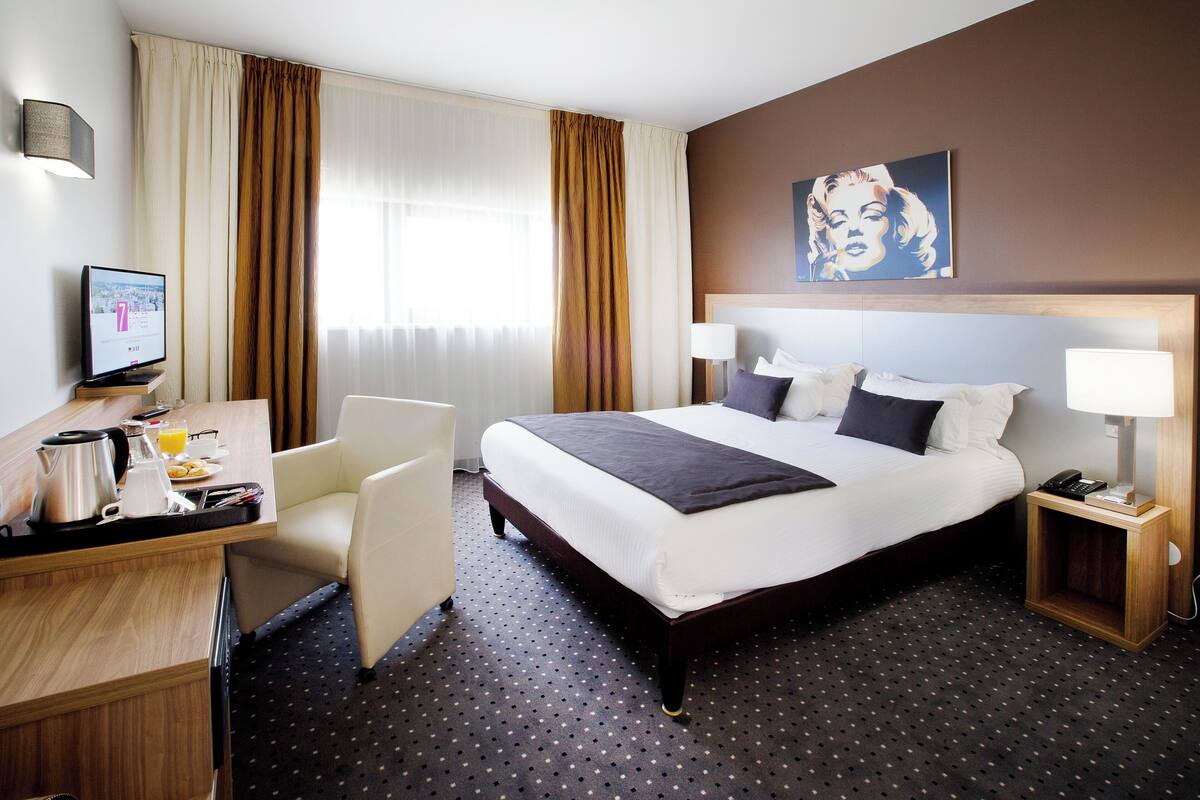 A hotel room with a bed, bedside tables, a desk with a computer, a chair, and a window with curtains.