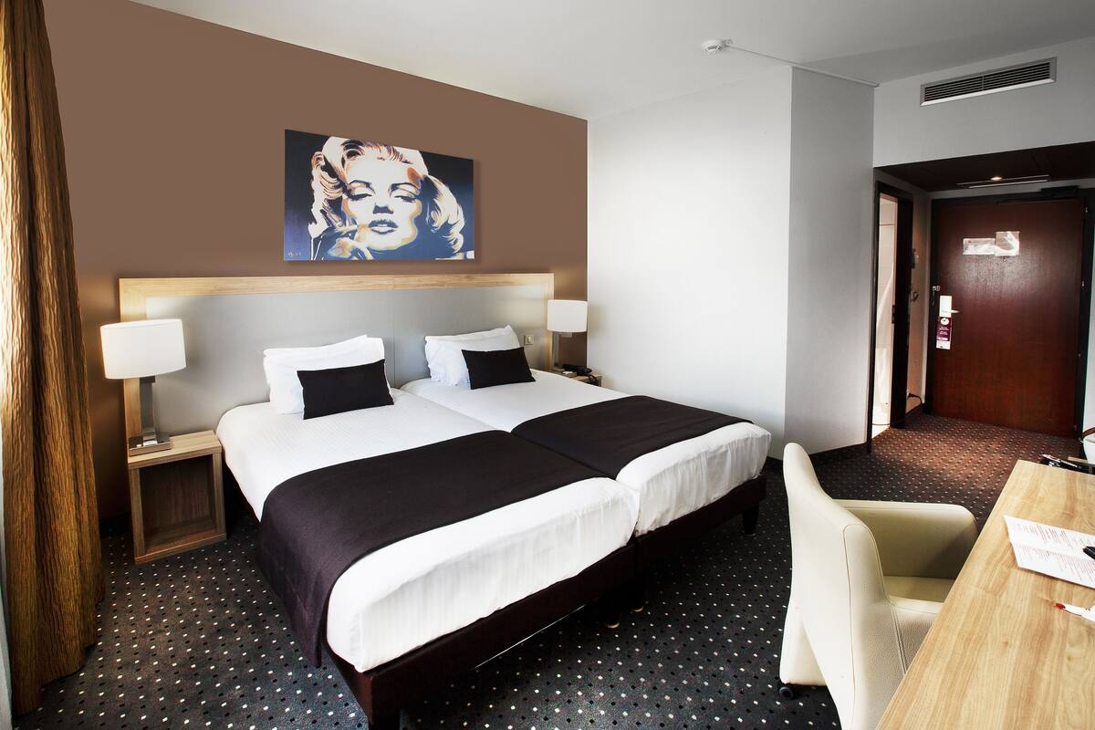 A hotel room with two beds, a desk, a chair, a lamp, and a painting on the wall.