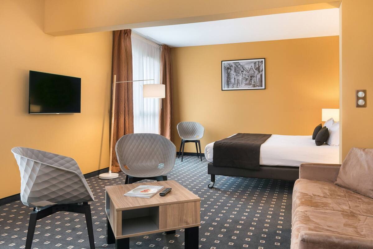 A hotel room with a bed, two chairs, a TV, a lamp, and a painting on the wall.