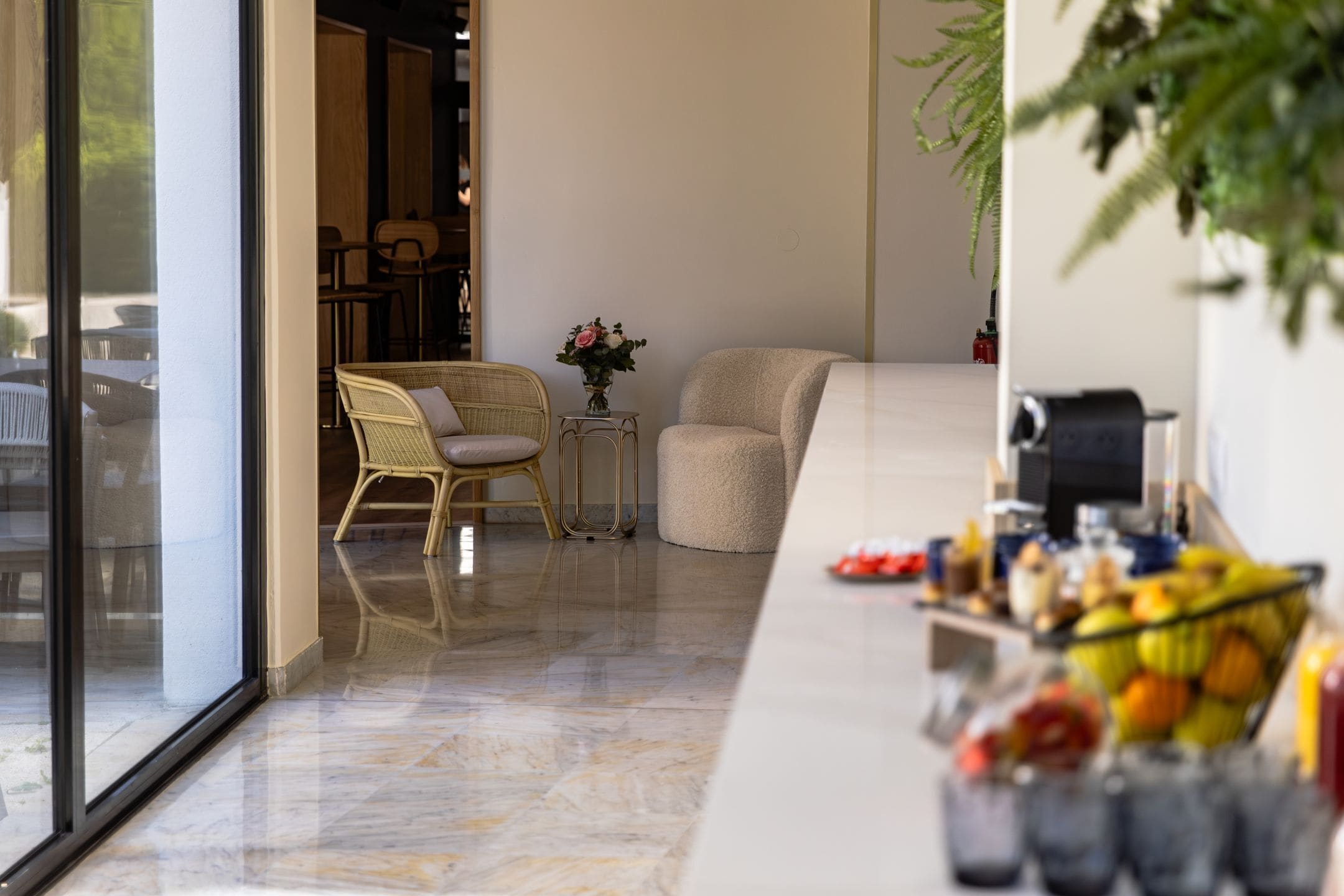 A modern hotel lobby with a glass door, a wicker chair, a small table with a vase of flowers, and a fruit basket on a counter.