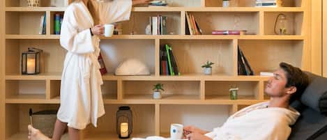 A room with a large wooden bookshelf, a black sofa, and a person in a bathrobe holding a mug.