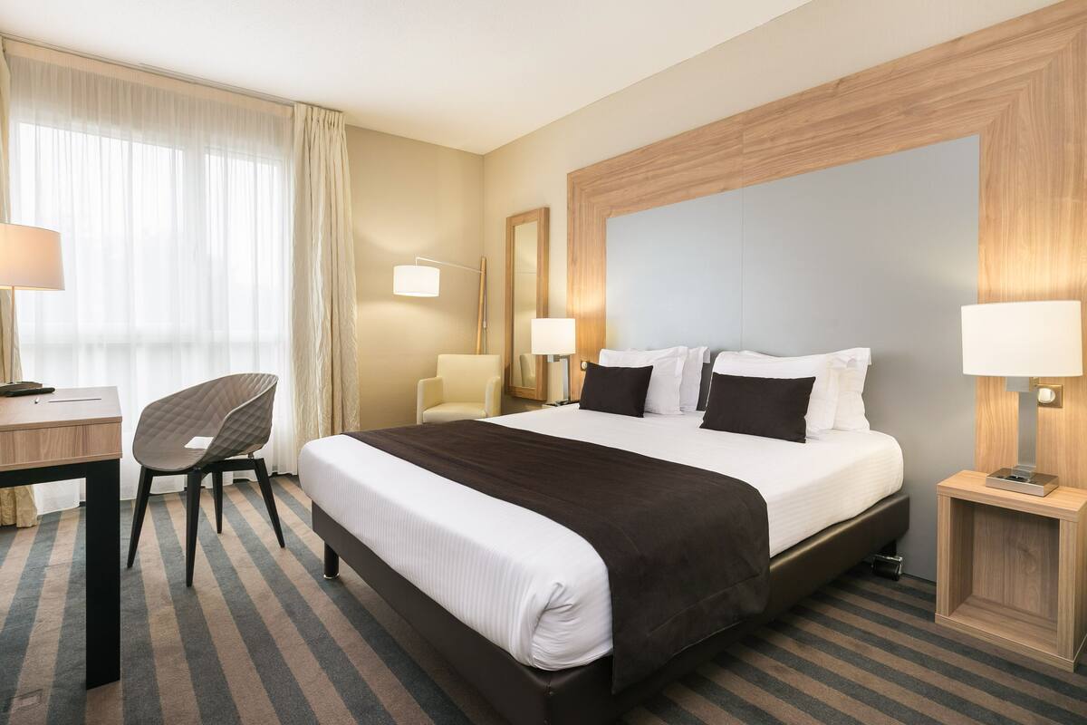 A hotel room with a large bed, a desk, a chair, and a nightstand with a lamp.