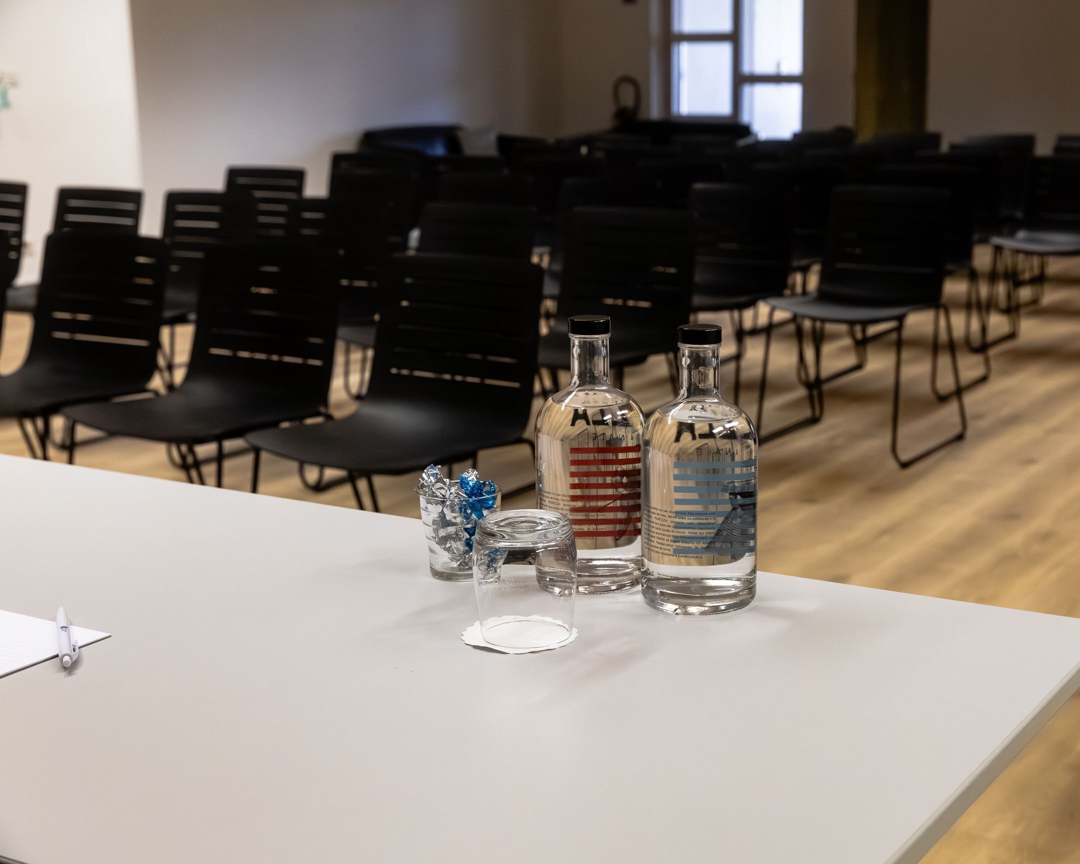 Un salón de conferencias con filas de sillas negras, una mesa blanca con tres botellas y un vaso, y una ventana al fondo.