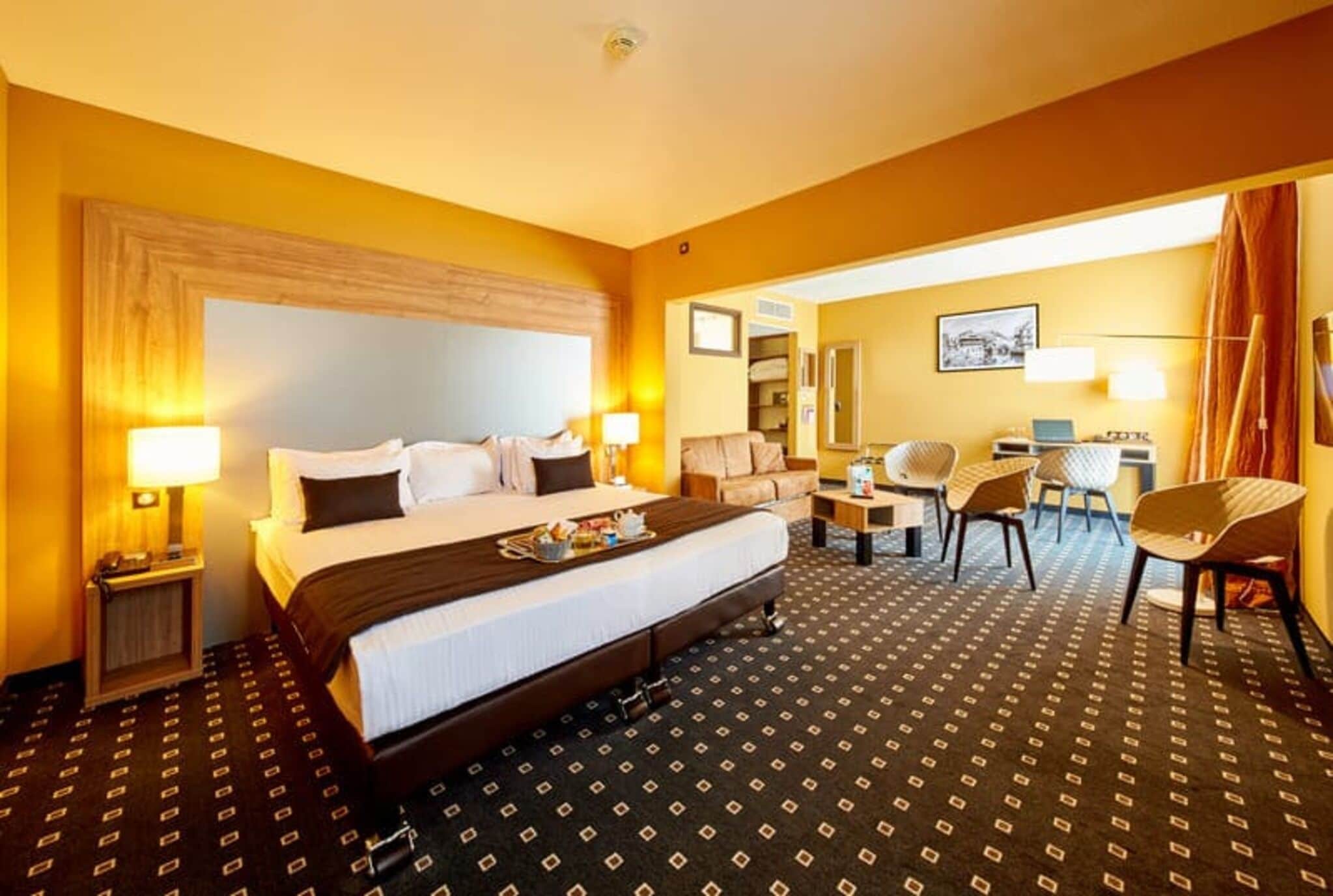 A hotel room with a large bed, a desk, two chairs, a small table, and a mirror.