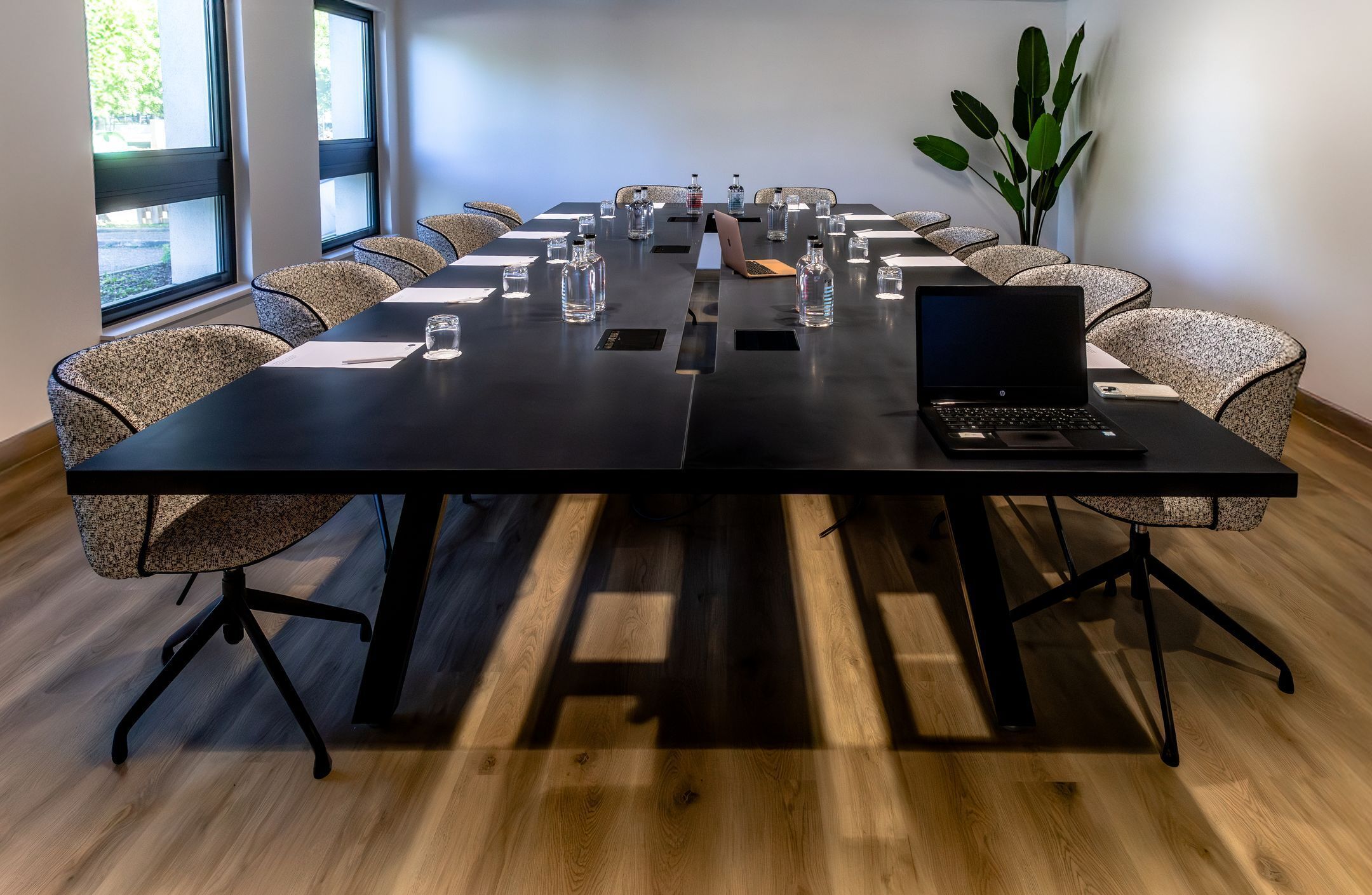 Un salón de conferencias con una mesa larga, sillas, botellas de agua, una computadora portátil y una planta.