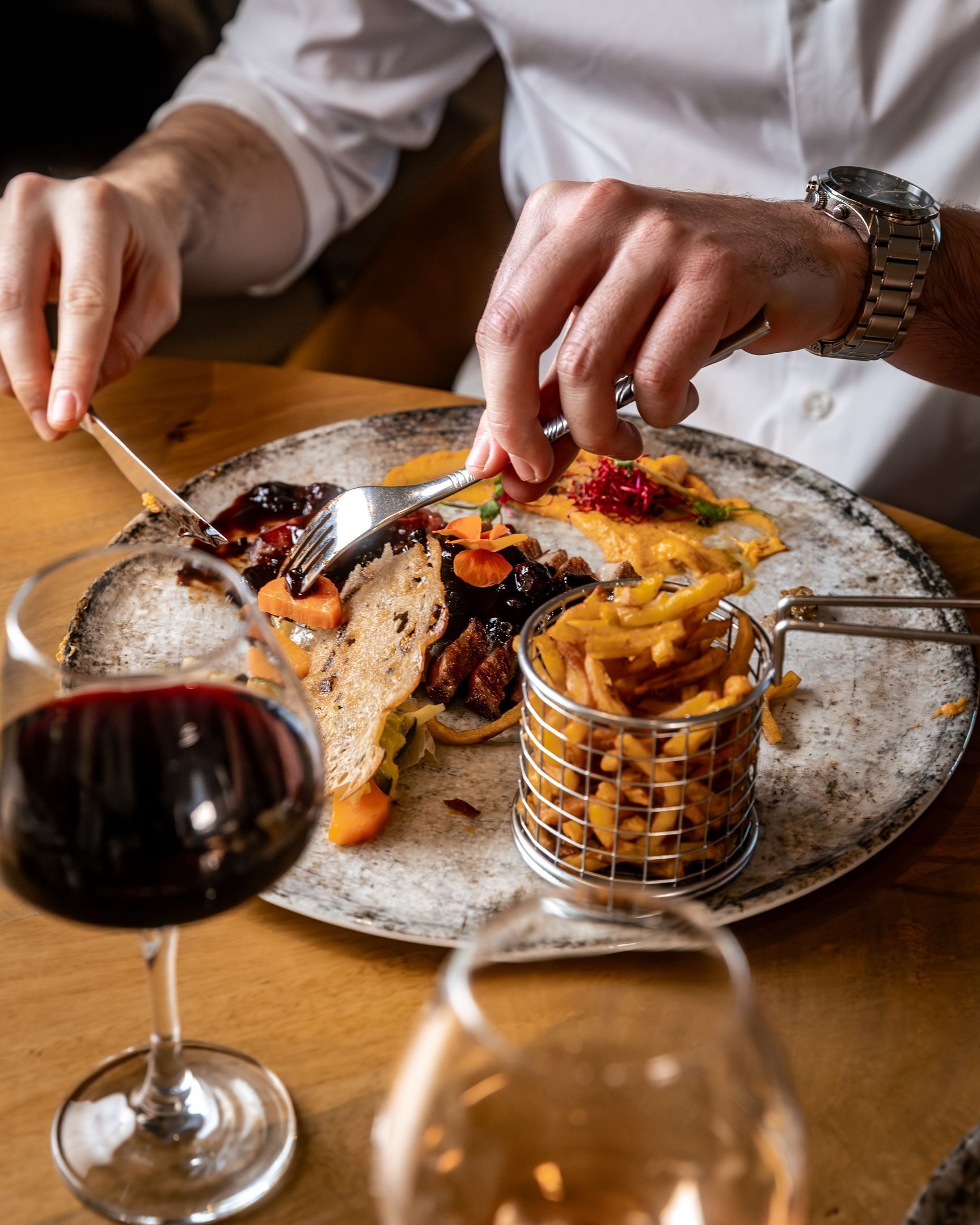 Eine Person isst an einem Tisch mit einem Teller Essen, einem Glas Rotwein und einer Metallbox mit Pommes.