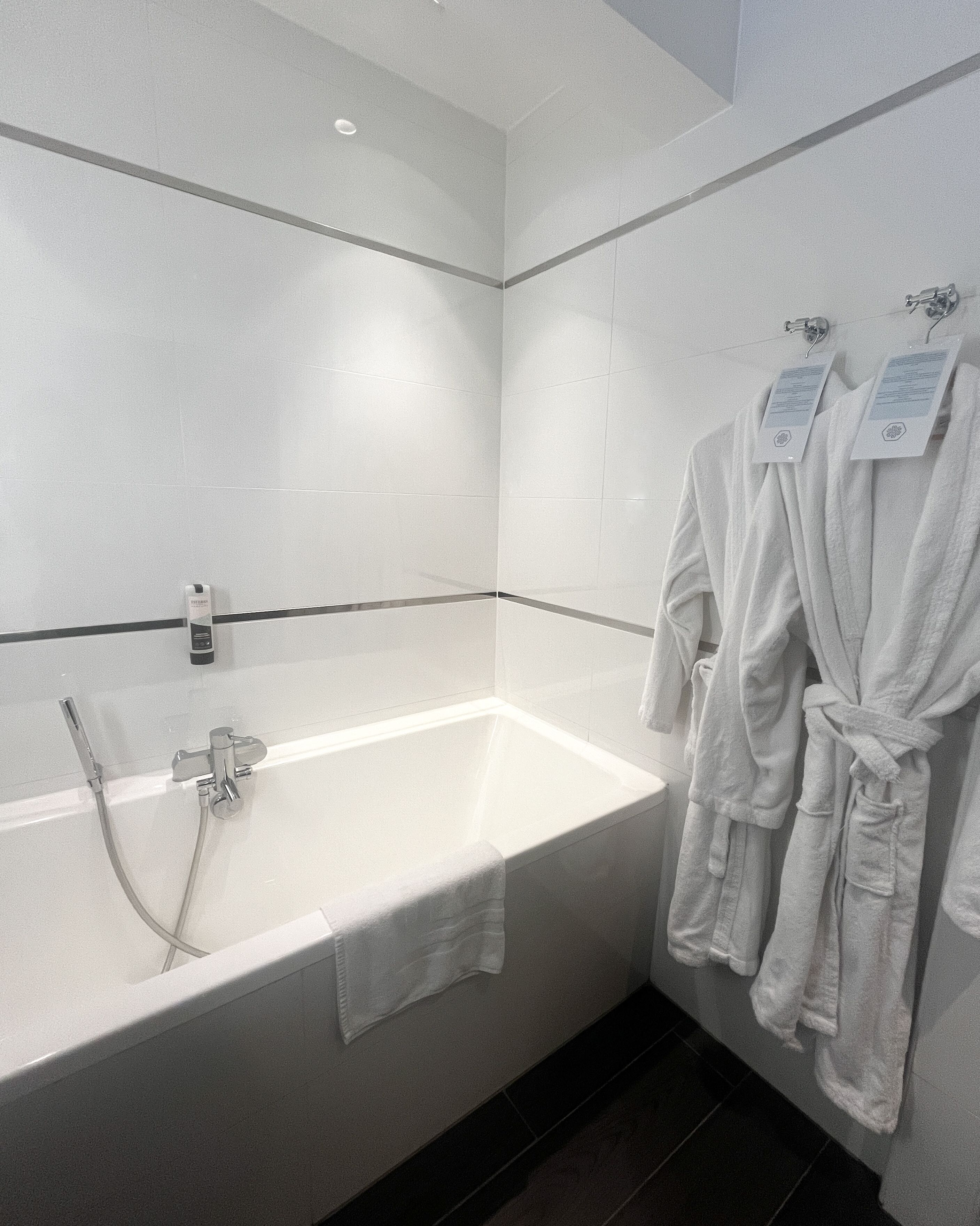 Une salle de bain moderne avec une baignoire blanche, une douchette et deux peignoirs blancs suspendus à des crochets.