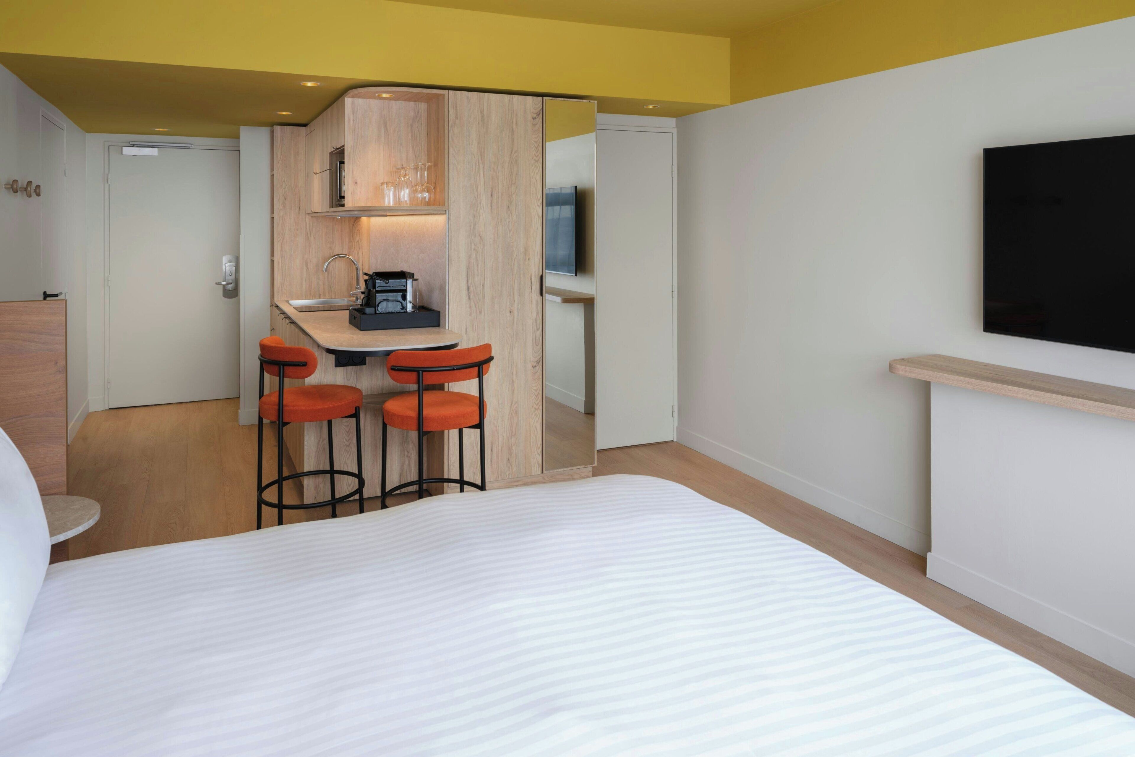 Une chambre d’hôtel comprenant un lit, une kitchenette avec une machine à café, deux tabourets de bar et un téléviseur fixé au mur.