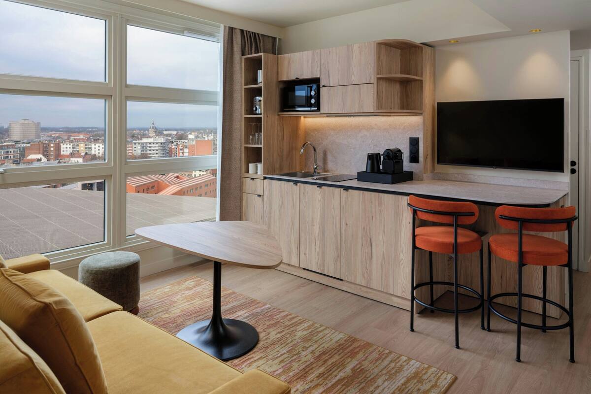 A modern kitchen with a large window, wooden cabinets, a flat-screen TV, and a bar area with stools.