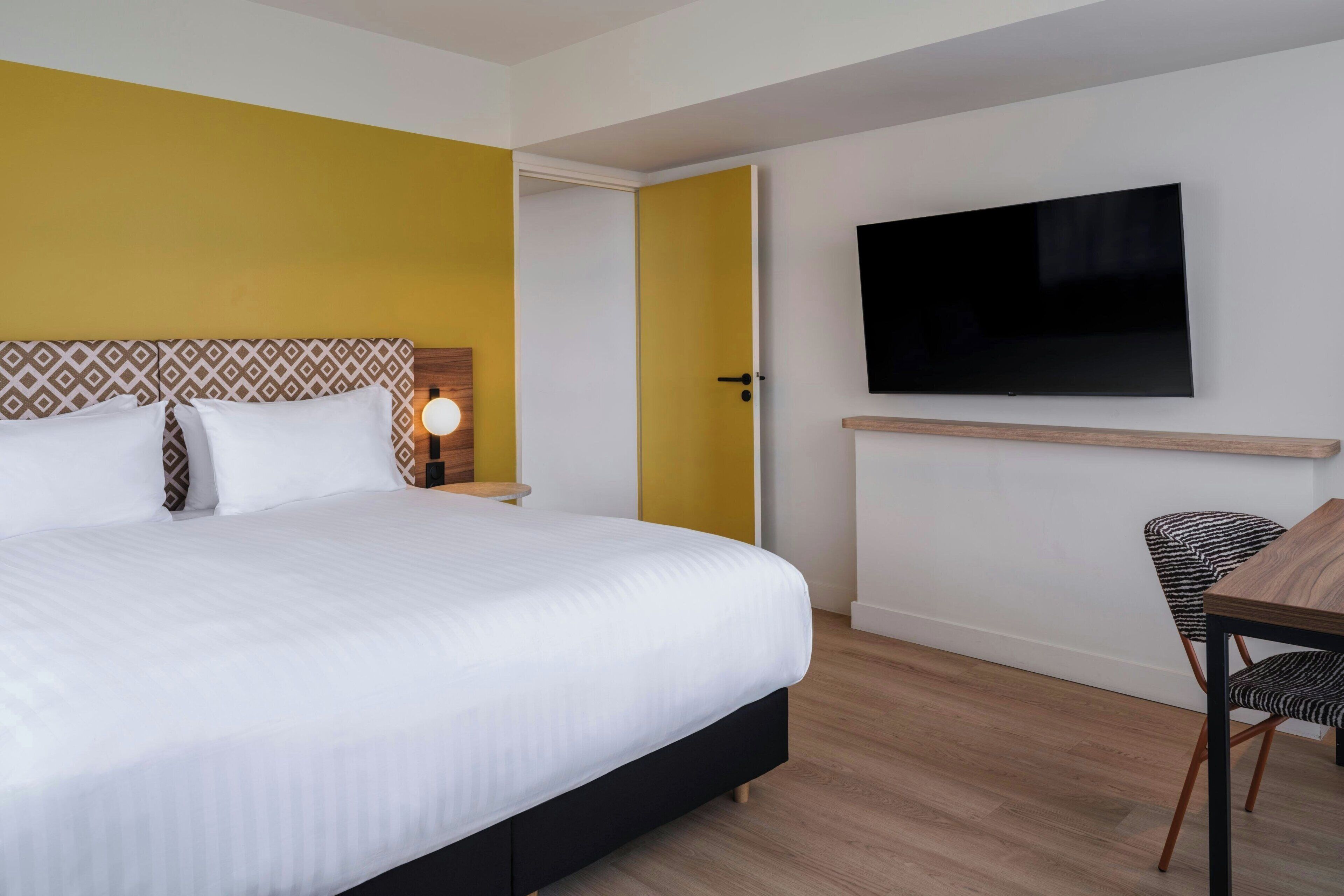 Una habitación de hotel con una cama grande, un televisor montado en la pared, una silla de madera y una mesita de noche con lámpara.