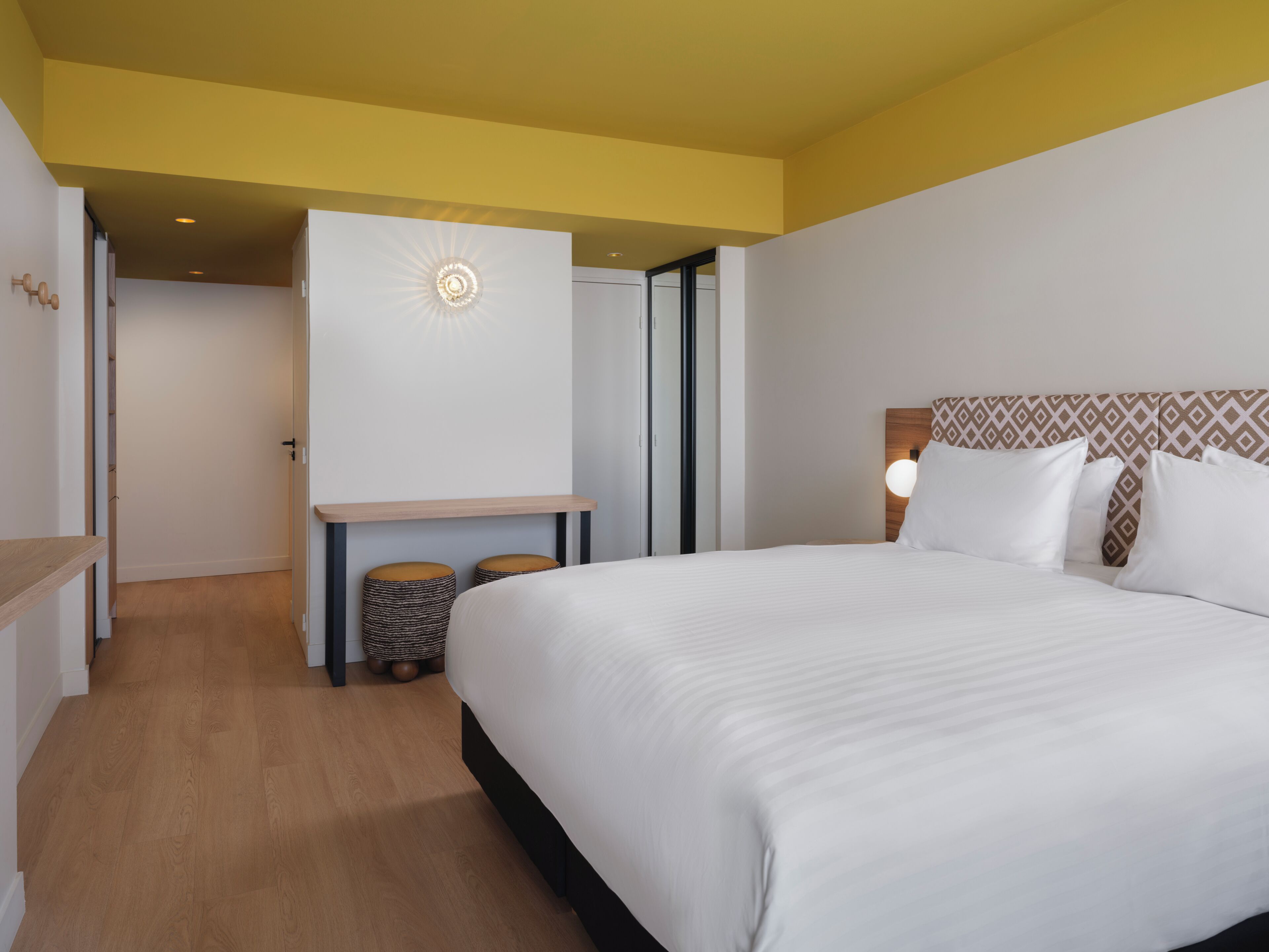 A hotel room with a bed, a desk, a chair, and a small stool.