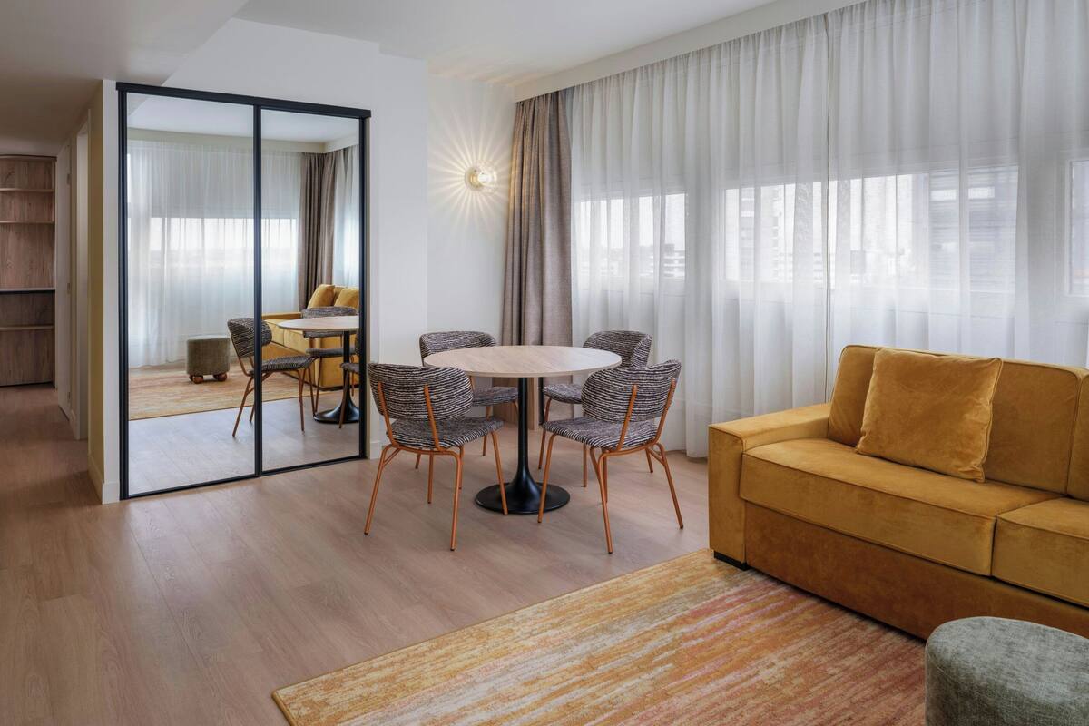 A modern living room with a round dining table, two sofas, and a large window with sheer curtains.