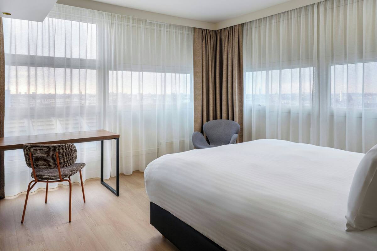 A hotel room with a large bed, a desk, a chair, and a view of the cityscape through sheer curtains.