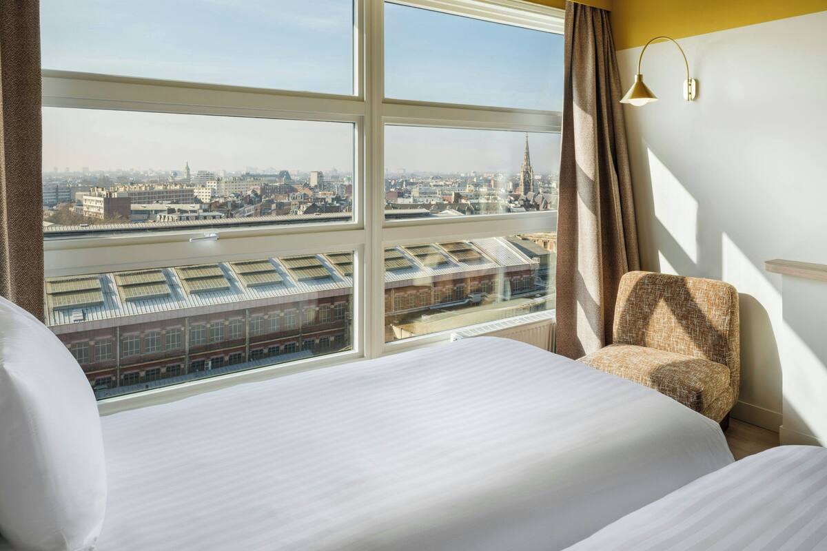 Una habitación de hotel con un ventanal grande, una cama, una silla y una lámpara.