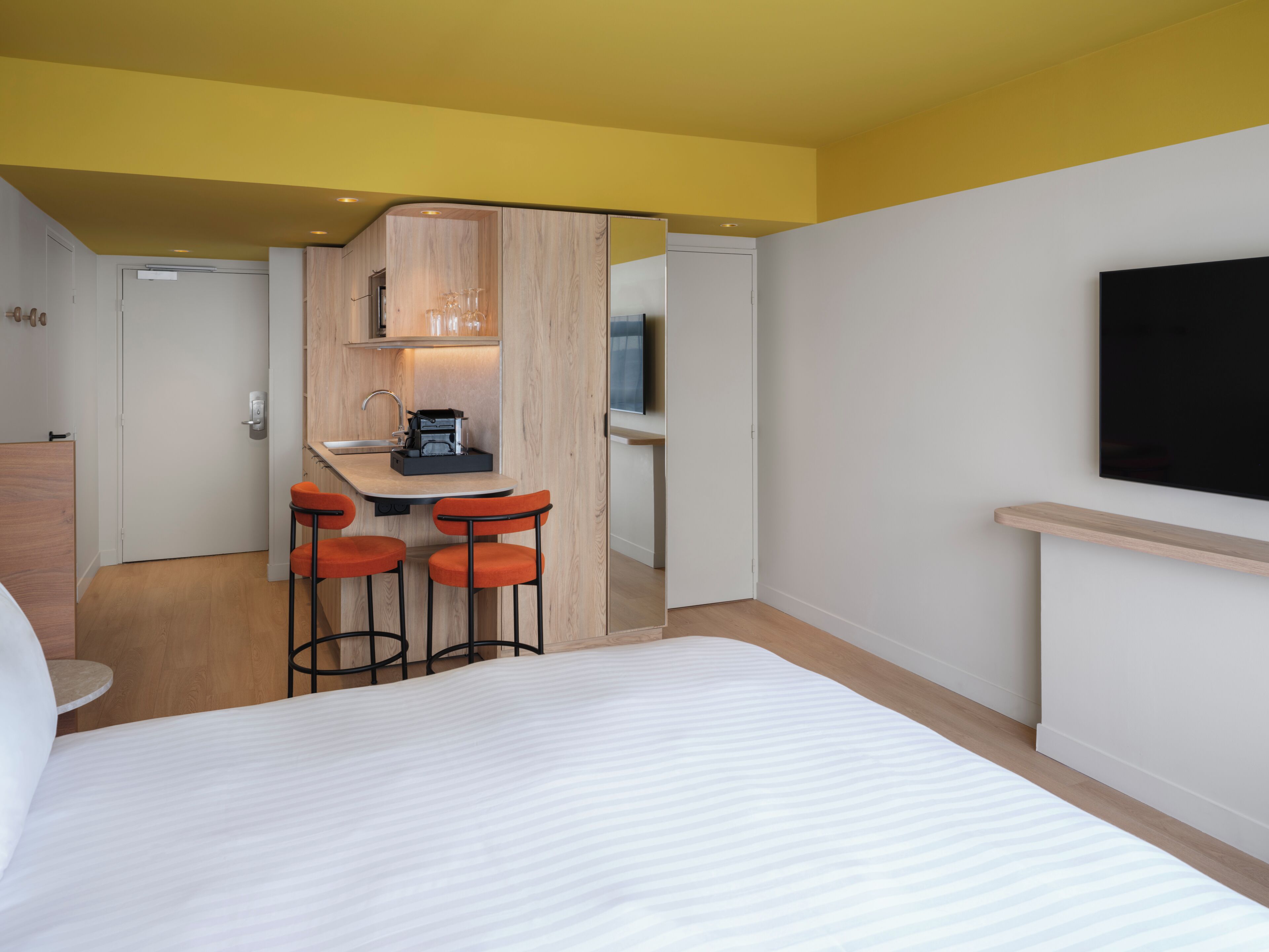 A hotel room with a bed, a small kitchenette, a TV, and a seating area with stools.