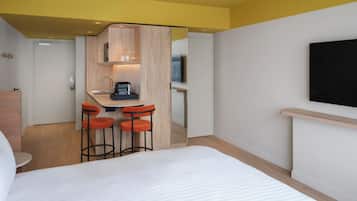 A hotel room with a bed, a small kitchenette, a TV, and a seating area with stools.