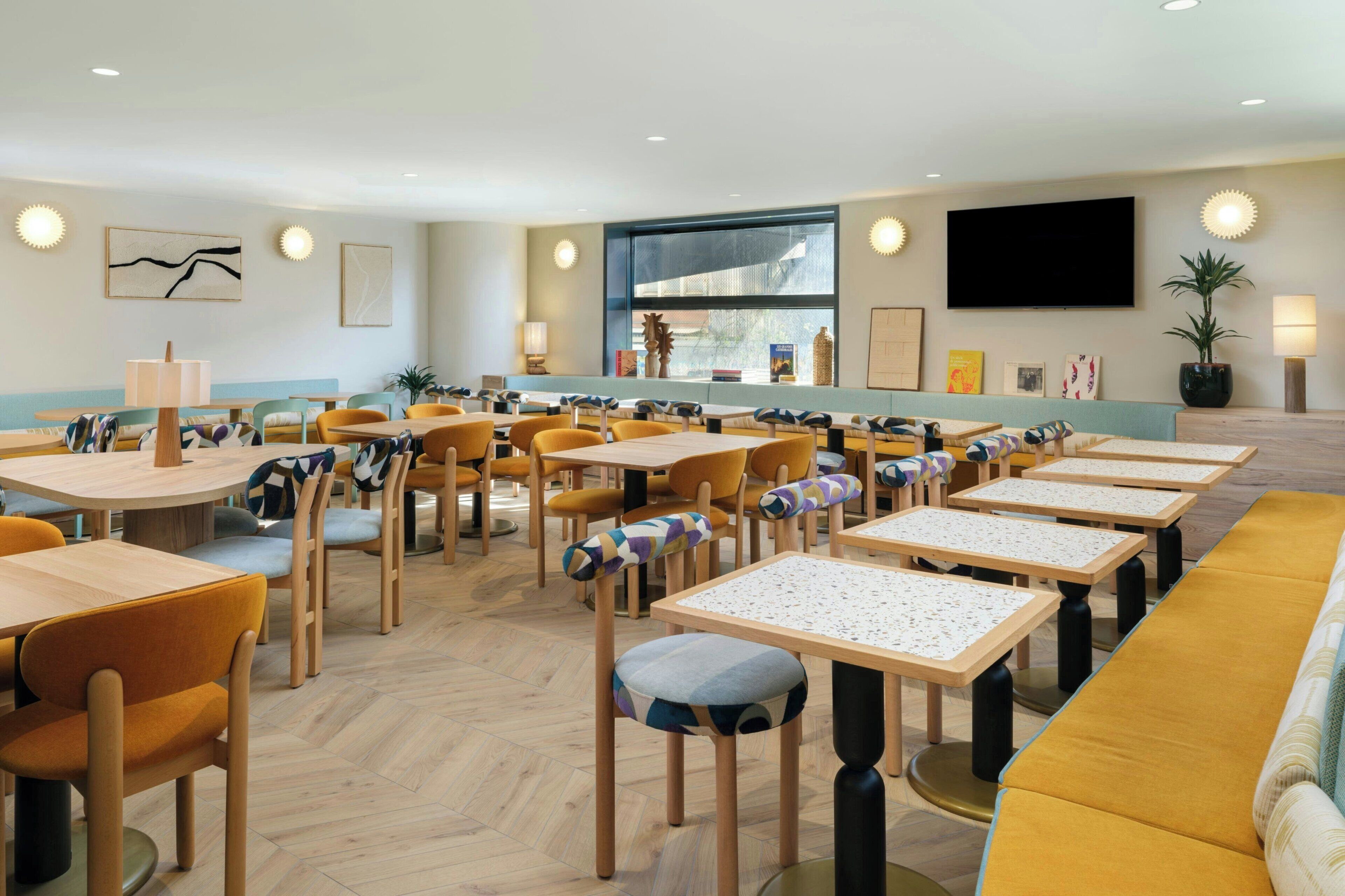 Un comedor moderno con mesas y sillas de madera, un sofá amarillo y un ventanal grande.