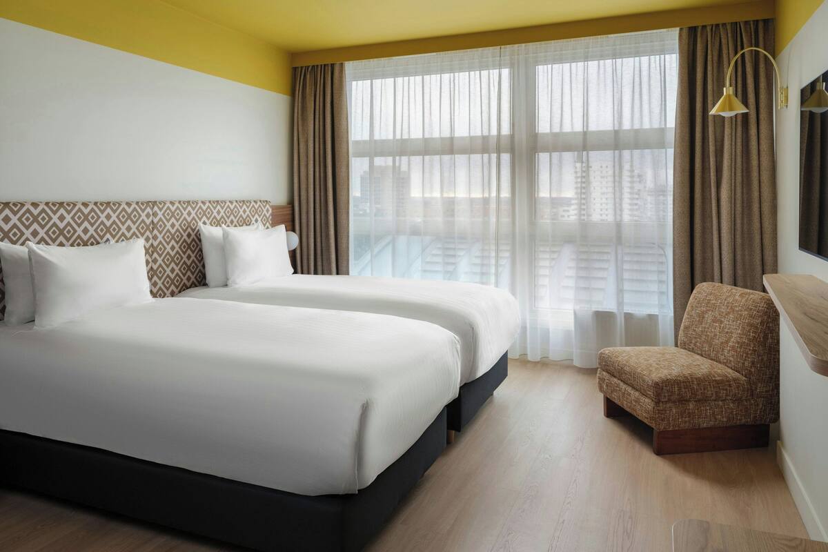 Habitación de hotel con dos camas, un sillón pequeño, un ventanal grande con cortinas y vistas a edificios.