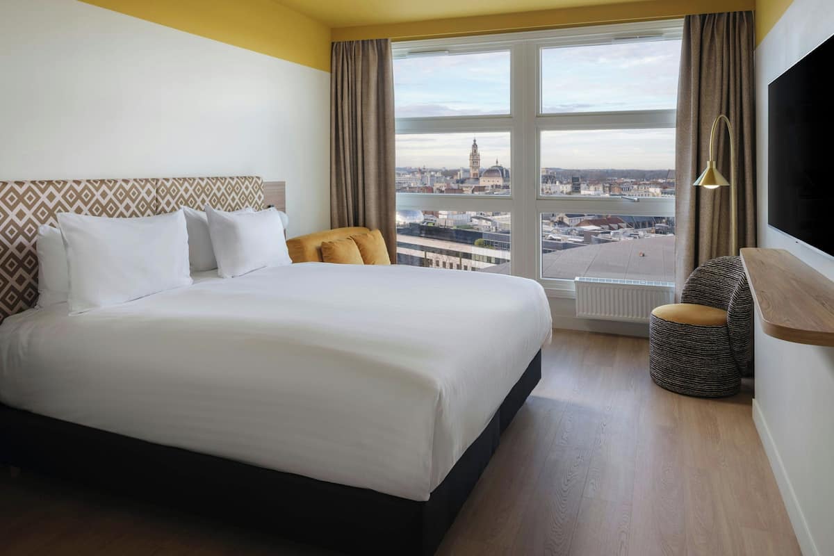 Habitación de hotel con una cama grande, vistas de la ciudad, televisión y ventana con cortinas.