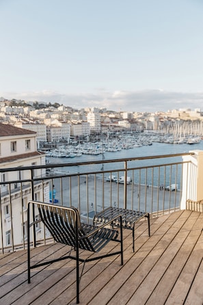 Un balcon avec deux chaises en métal et une terrasse en bois donnant sur une marina, avec des bateaux et des bâtiments en arrière-plan.