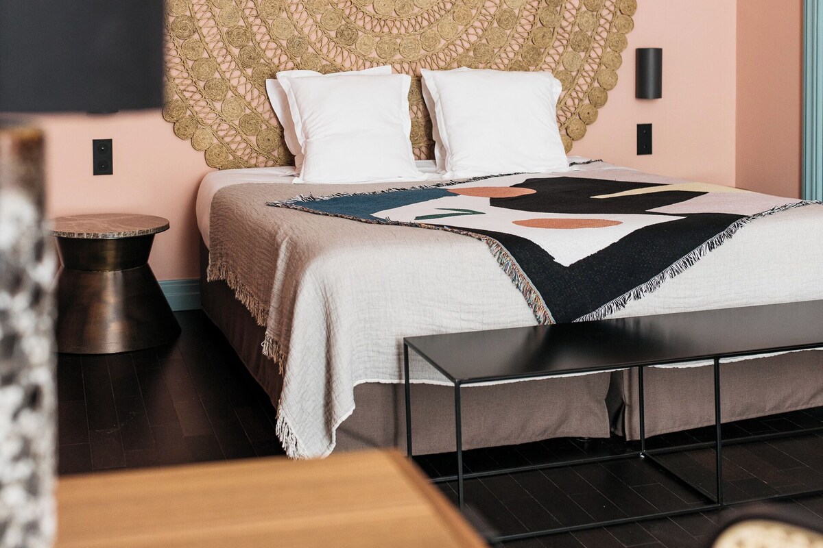 Un dormitorio con un cabecero grande tejido, una cama con ropa blanca, una mesita de noche y una lámpara.