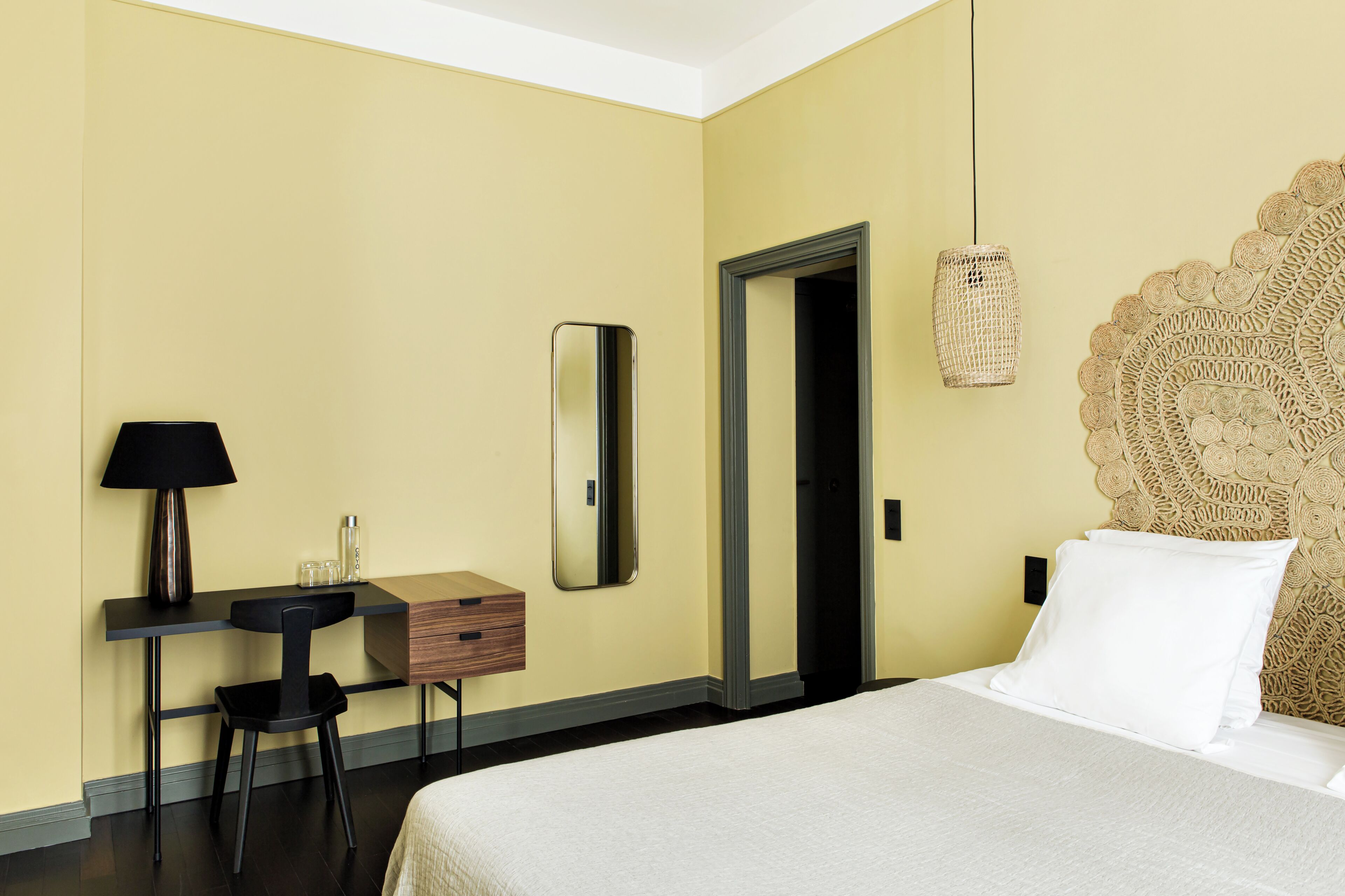 Een hotelkamer met een bed, een bureau met een lamp, een spiegel en een wanddecoratie van geweven materiaal.