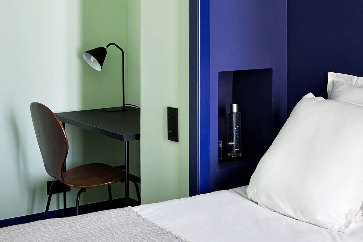 A bedroom with a bed, a desk with a chair, a lamp, and a bottle on the nightstand.