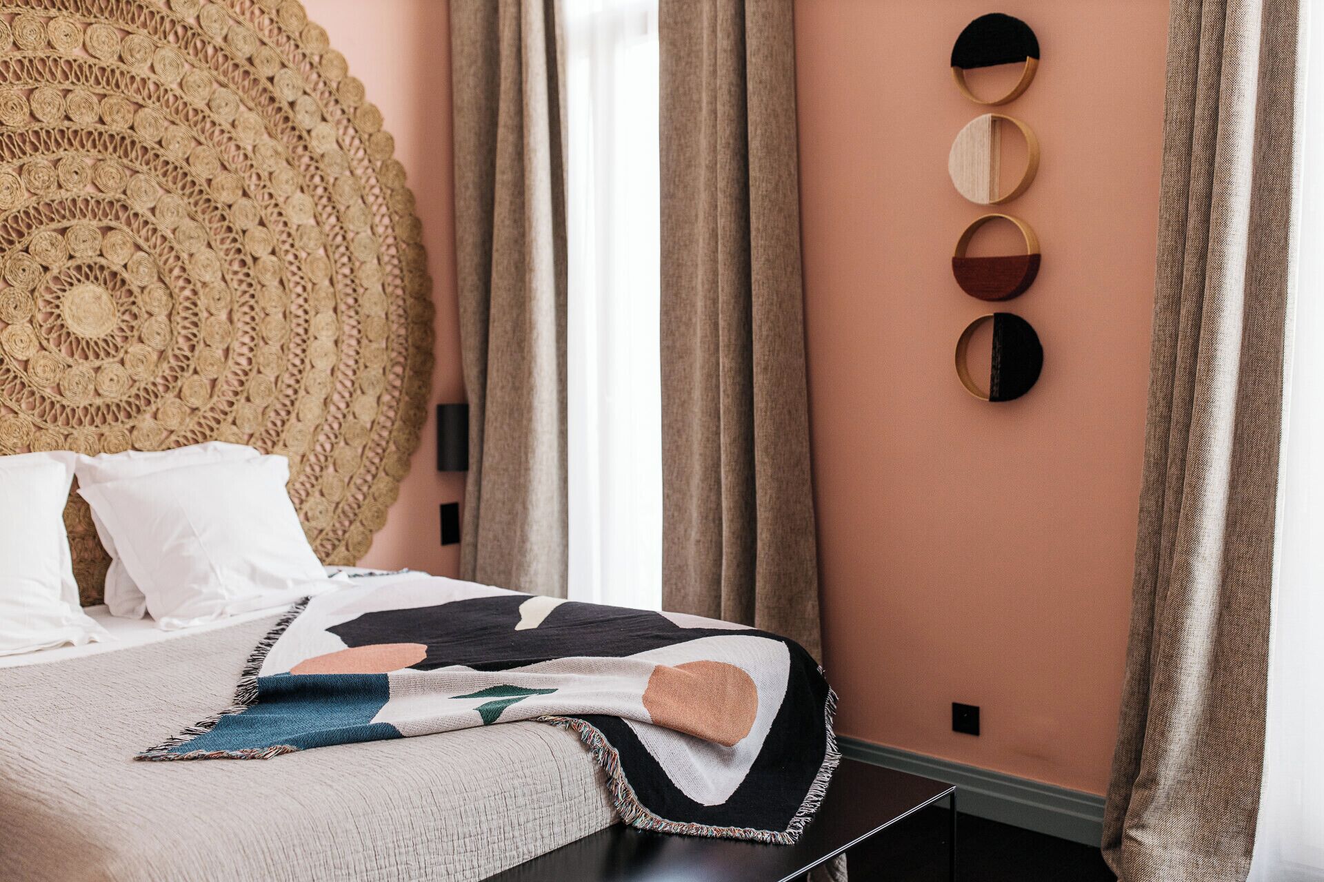 Un dormitorio con cama, un cabecero grande tejido, un espejo montado en la pared y una ventana con cortinas.