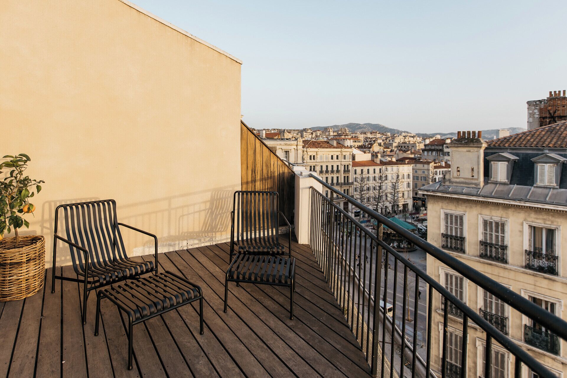 A balcony with two chairs and a wicker basket, overlooking a cityscape with buildings.