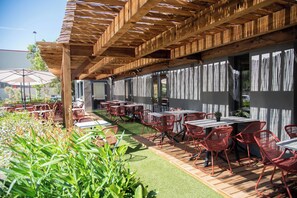 Une terrasse extérieure avec des tables en bois et des chaises rouges, entourée de verdure et agrémentée d’une pergola en bois.