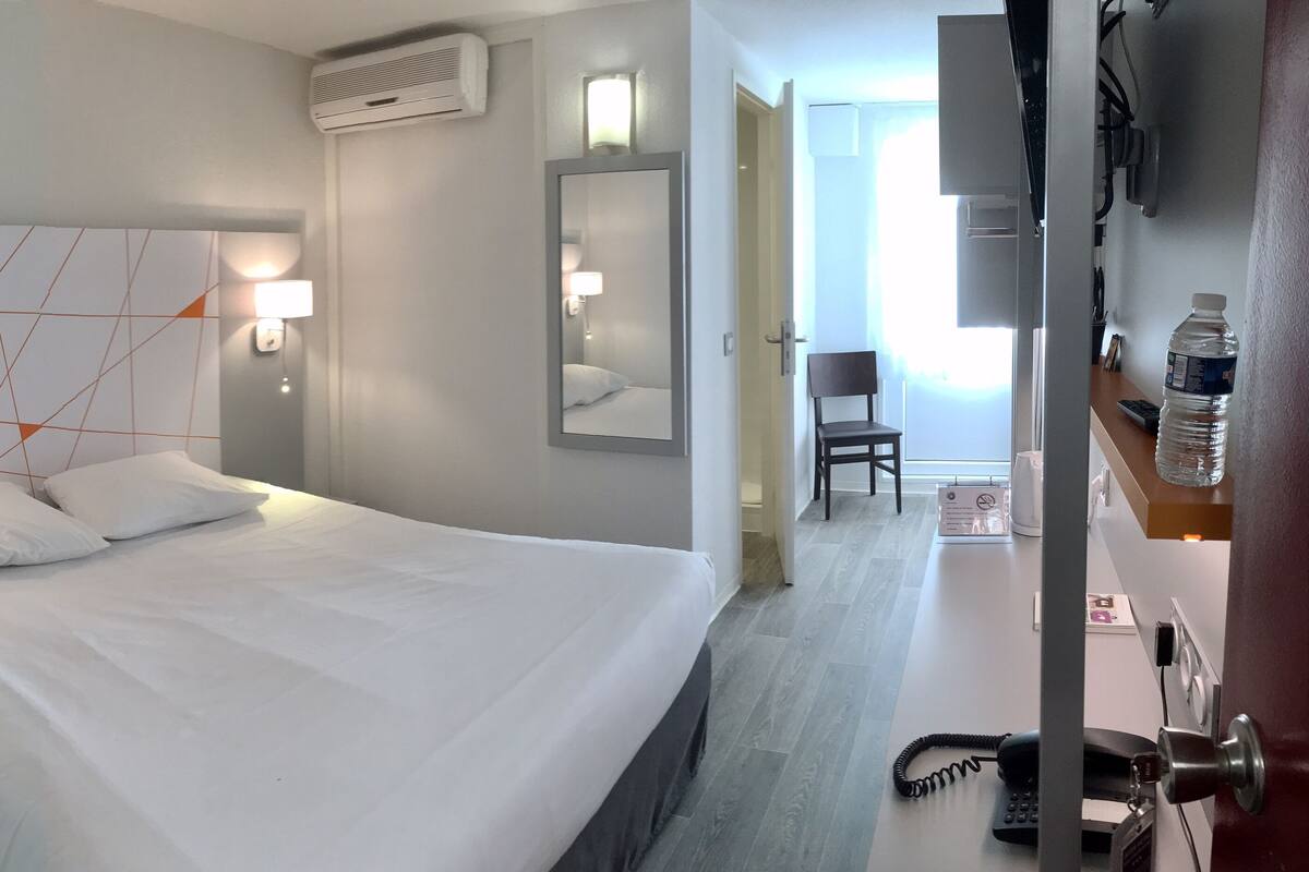 Ein Hotelzimmer mit einem großen Bett, Nachttischlampen, einem Spiegel, einem Schreibtisch mit Stuhl und einer Wasserflasche.
