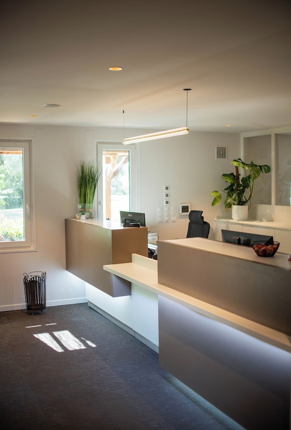 A modern reception area with a desk, computer, and seating. There are plants and a window providing natural light. - Hôtel du Parc Euromédecine (Grabels)