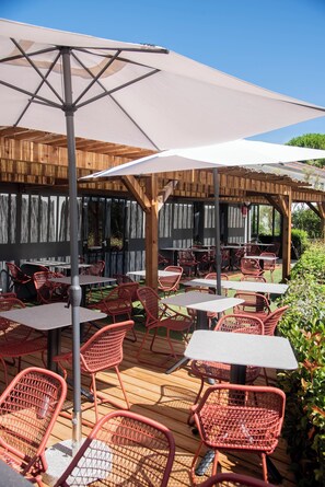 Une terrasse extérieure avec des chaises et des tables en osier rouge, une pergola surmontée d’une toile blanche et une plateforme en bois.