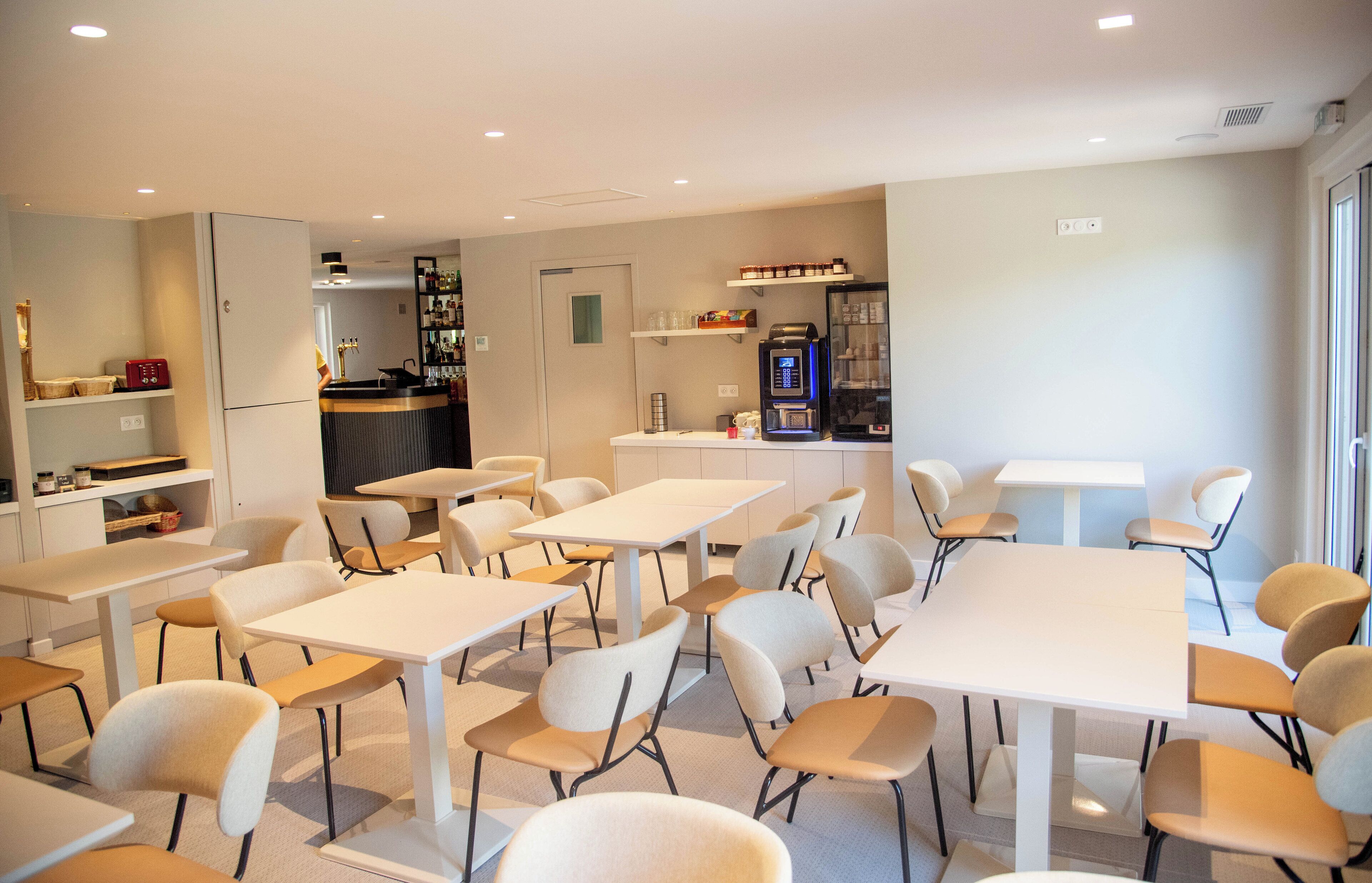 A modern dining area with white tables and chairs, a kitchen area with a coffee machine, and a counter with various items.
