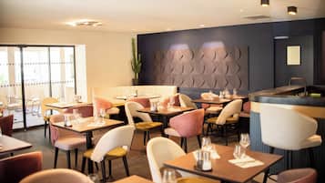 Een restaurant met meerdere tafels en stoelen, waarvan sommige voorzien zijn van kussens. Er staan planten en er is een decoratieve wand.