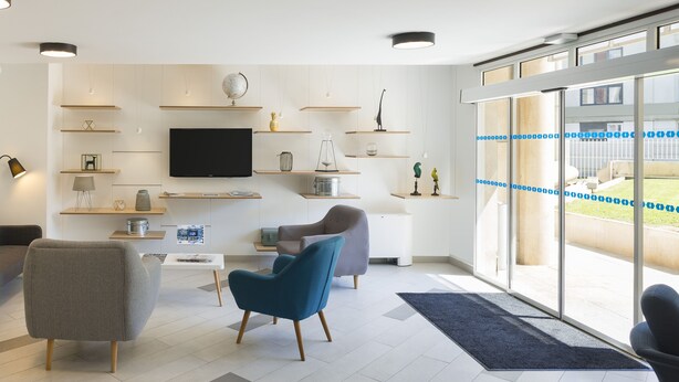 Una sala de estar moderna con un televisor de pantalla plana, dos sofás y una mesa de centro. Se ven estanterías con objetos decorativos y un ventanal con vistas al exterior.