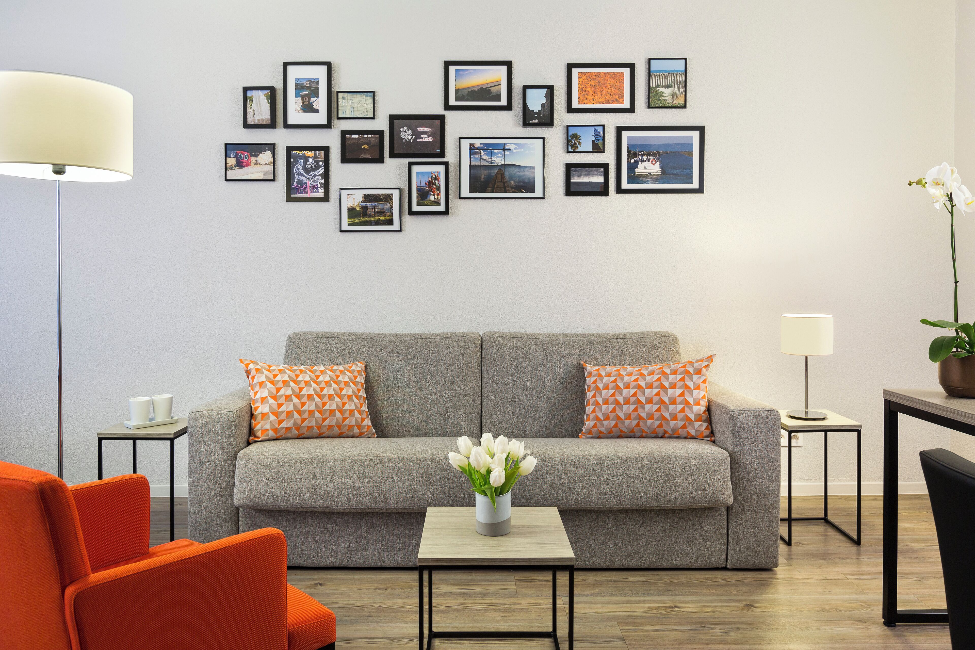Una sala de estar moderna con un sofá gris, almohadas naranjas, una mesita con un jarrón de flores y una pared decorada con fotografías enmarcadas.